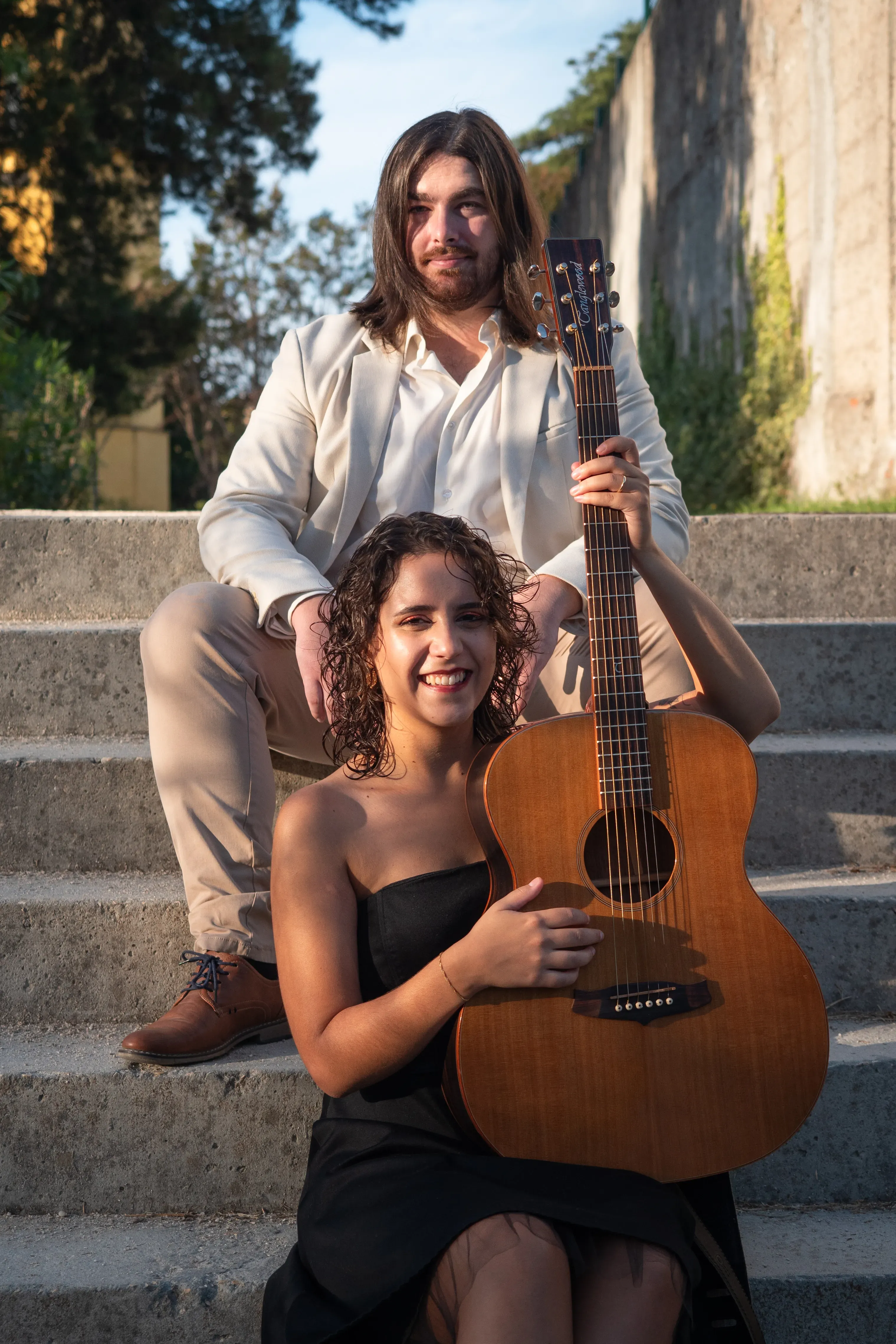 Numa escadas de betão de um parque, um casal está sentado. O rapaz está em cima, tem cabelo comprido liso e está vestido com um fato claro. A rapariga, mais em baixo, está a segurar a guitarra clássica na vertical e está vestida com um vestido preto e tem cabelo encaracolado curto.